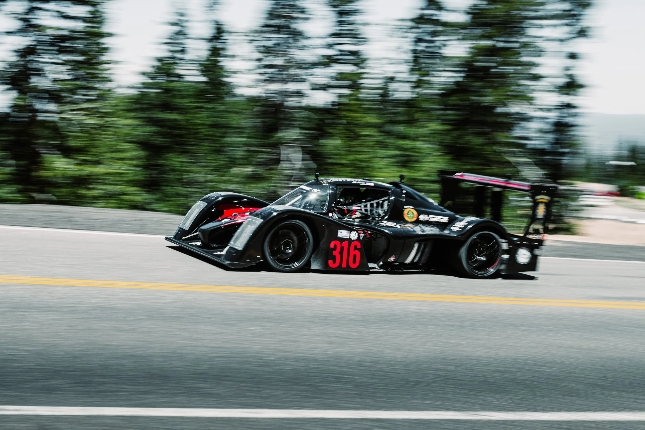 A black race car with the number 316 driving on a road surrounded by trees, appearing blurred due to speed.