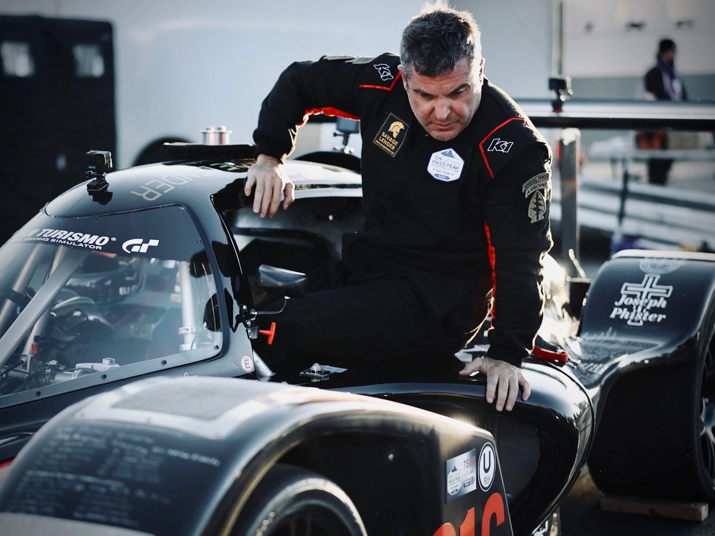 A man in a black racing suit getting out of the vehicle, with other people and race equipment in the background.