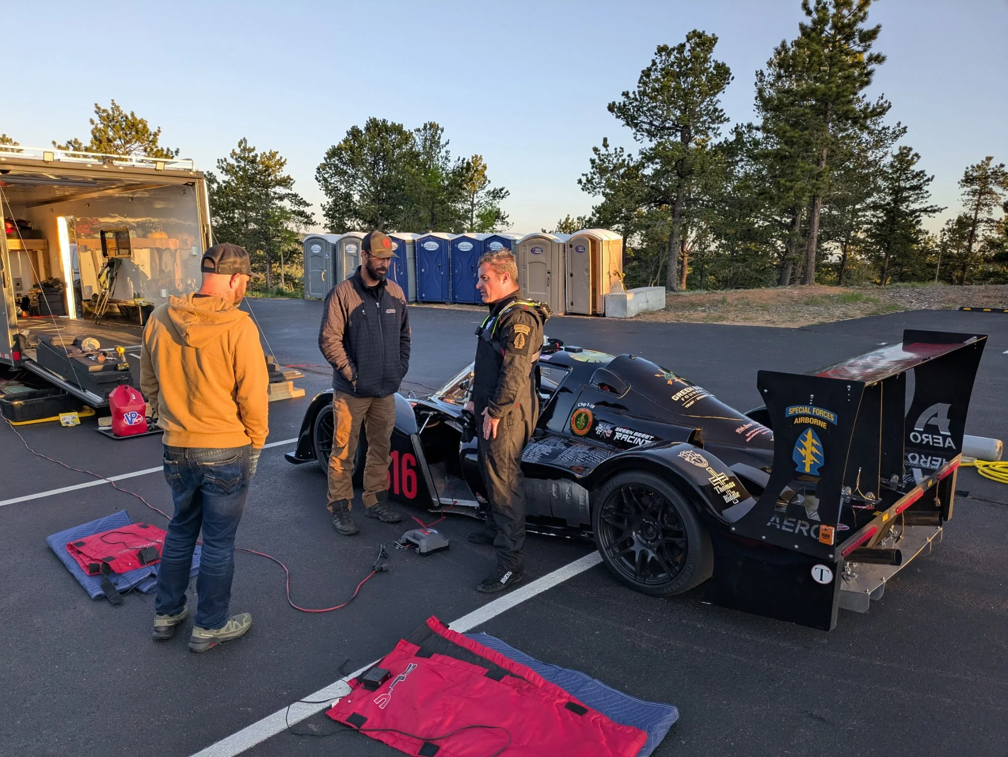 Three men standing beside a race car in a parking lot, with equipment laid out on the ground, and a row of portable toilets in the background.