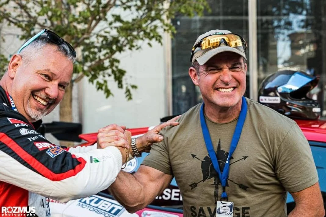Two men smiling and shaking hands, one dressed in a racing suit and the other in a t-shirt, outdoors with a car in the background.