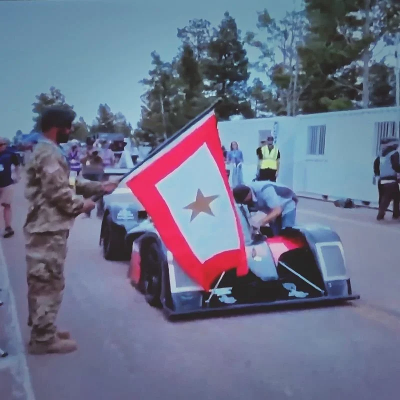 A person in military attire holding a red and white flag with a star near a race car, with people and trees in the background.