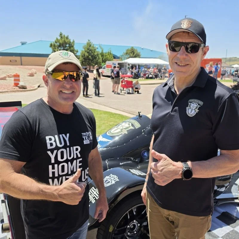 Two men smiling and giving thumbs up at an outdoor event with a black vehicle in the background.
