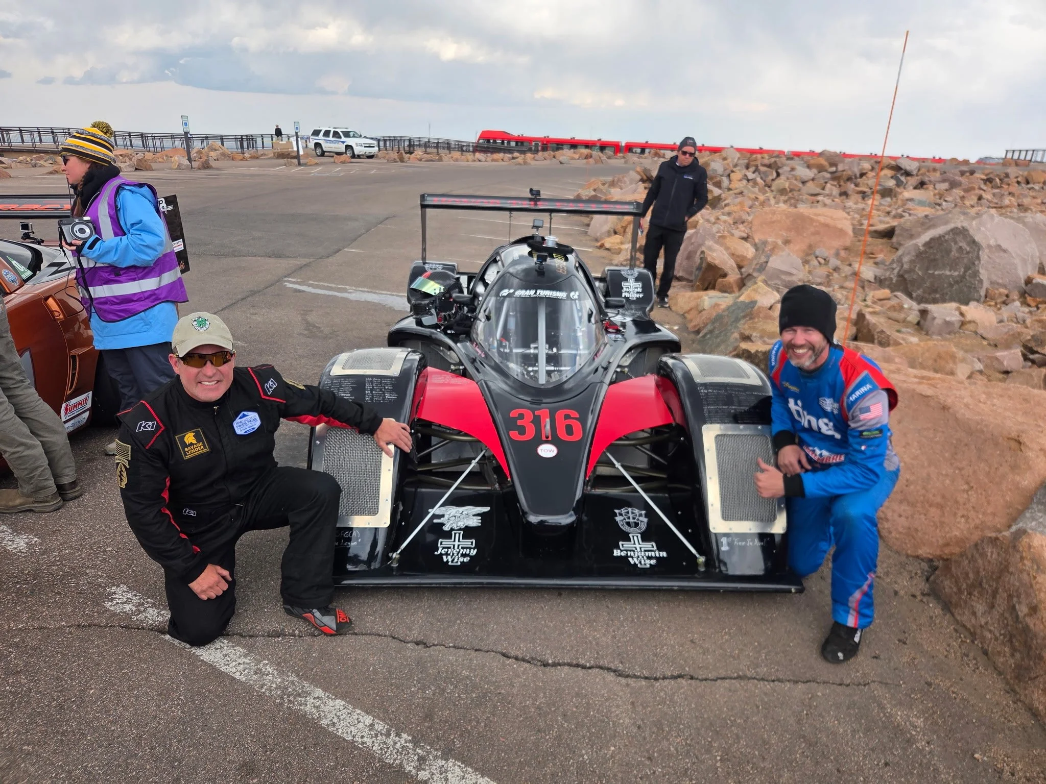 People gathered around a black and red race car with the number 316, in an outdoor parking lot with rocks and a cloudy sky in the background.