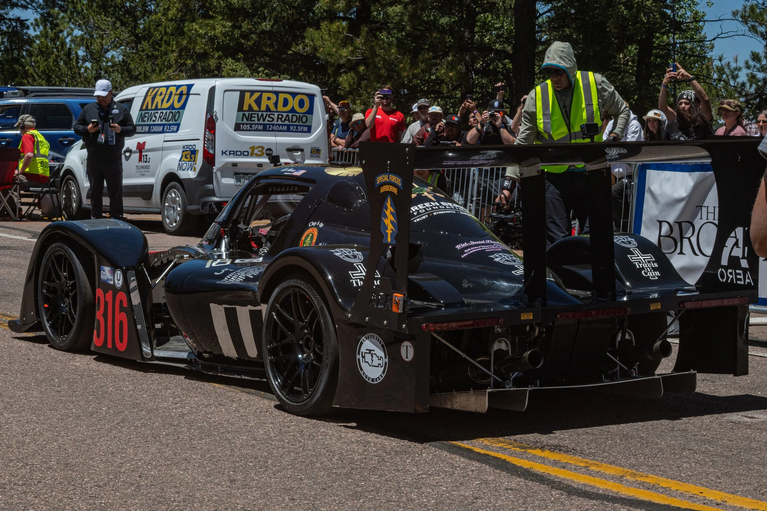 Black race car with number 316 on the front, surrounded by people and media at a racing event. Some individuals are taking photos, others are working or standing nearby, with trees in the background.
