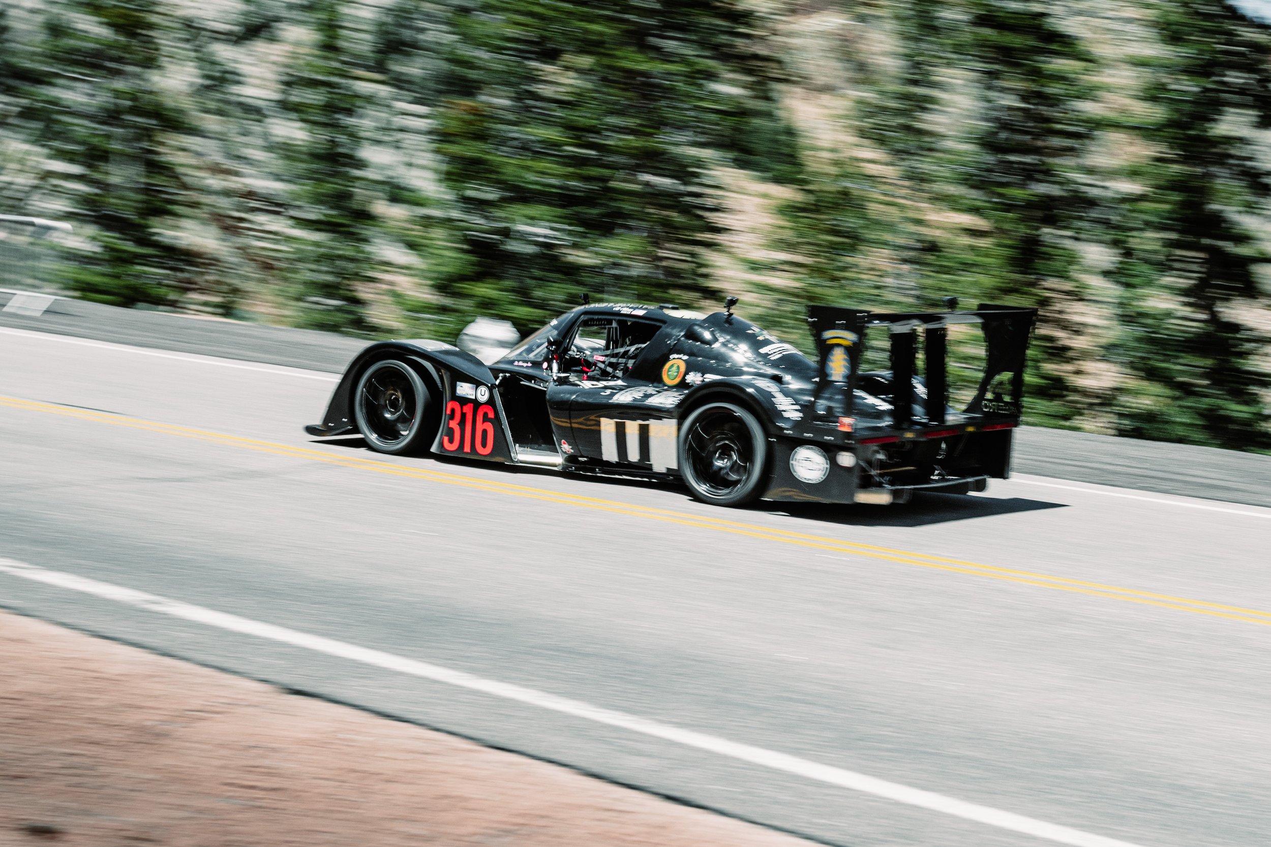 A sleek black racing car with the number 316 on the side speeding down a road through a lush green forest.