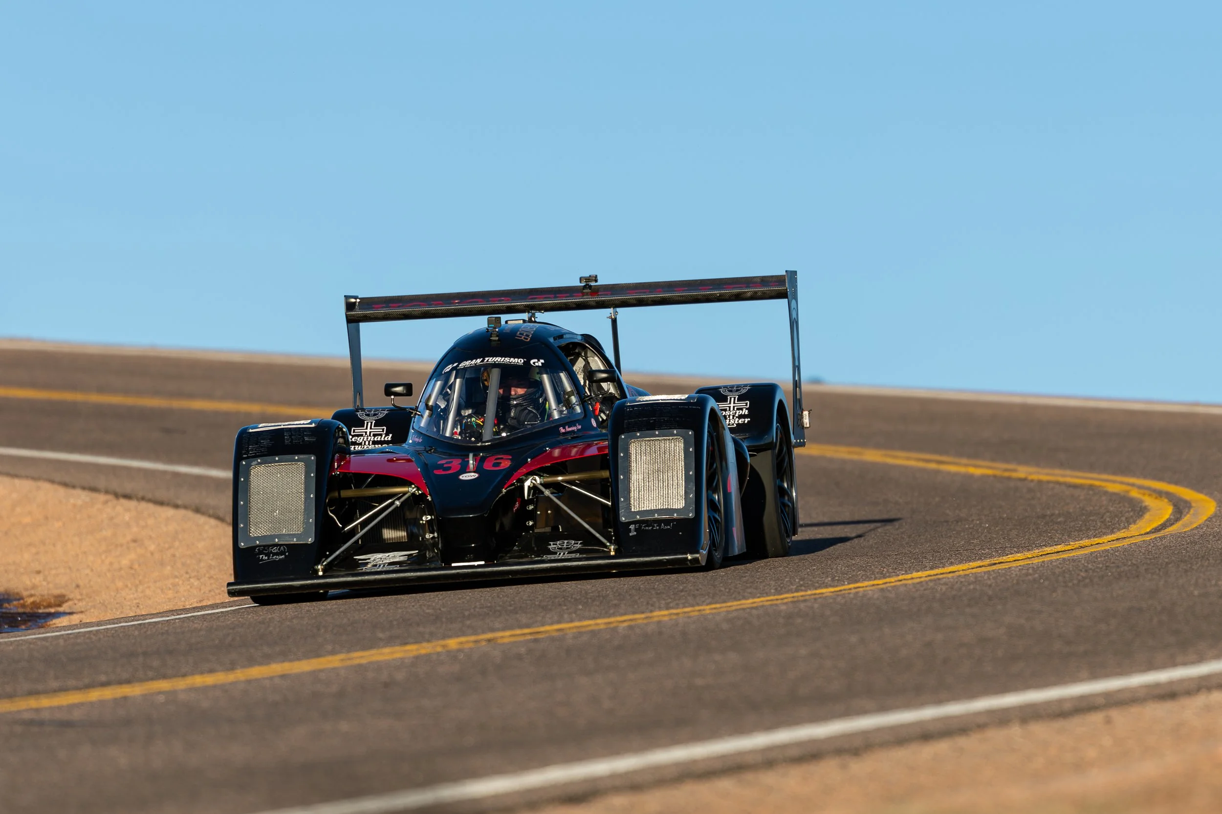 A black and red race car with the number 316 on the front is on a paved road with yellow lines, under a clear blue sky.