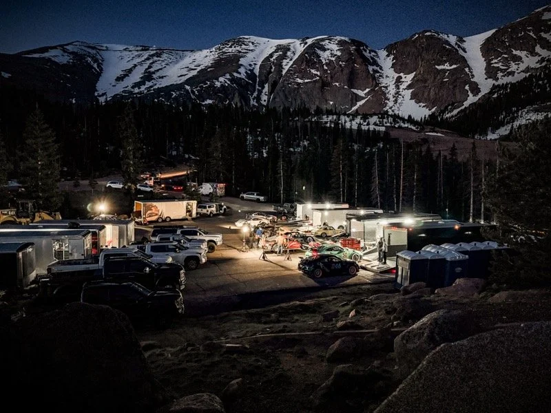 A parking lot at night with numerous parked vehicles, some trailers, and a few people. The background has snow-capped mountains under a dark sky.