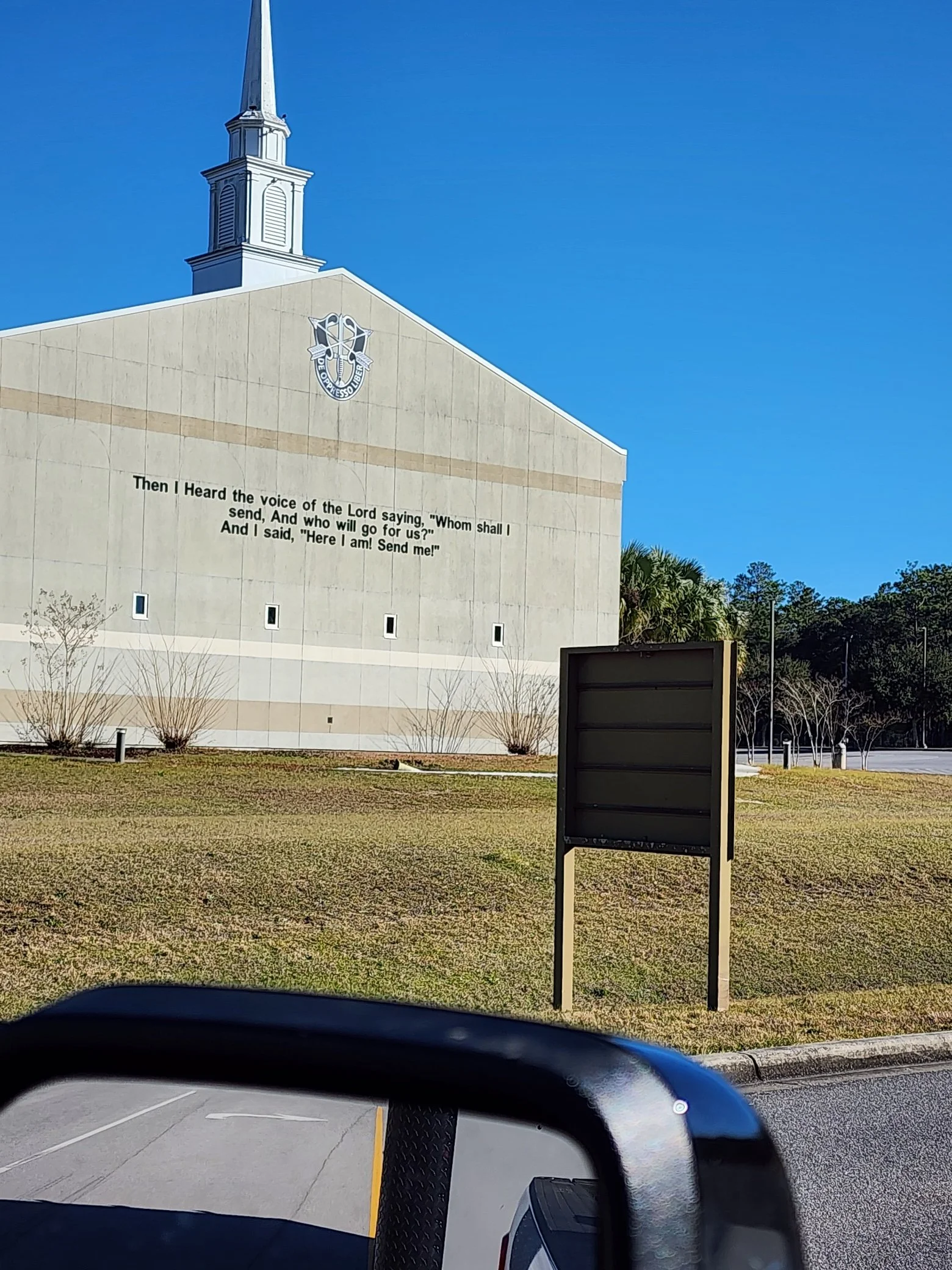 The outside of a church building with a tall steeple and a quote from the Bible on the wall that reads, 'Then I heard the voice of the Lord saying,