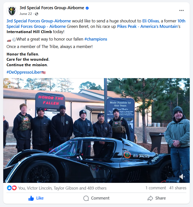 A group of seven men standing beside a black race car with a canopy, in front of a building. Two signs include one with pink text saying 'Honor the Fallen' and another with white text saying 'Made Possible by this Team." Some men wear motorcycle vest