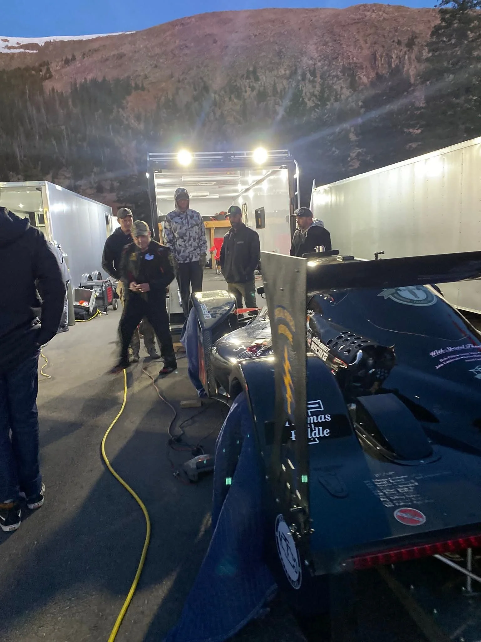 Group of people in racing team gear standing around race cars and equipment outdoors at dusk in mountainous area.