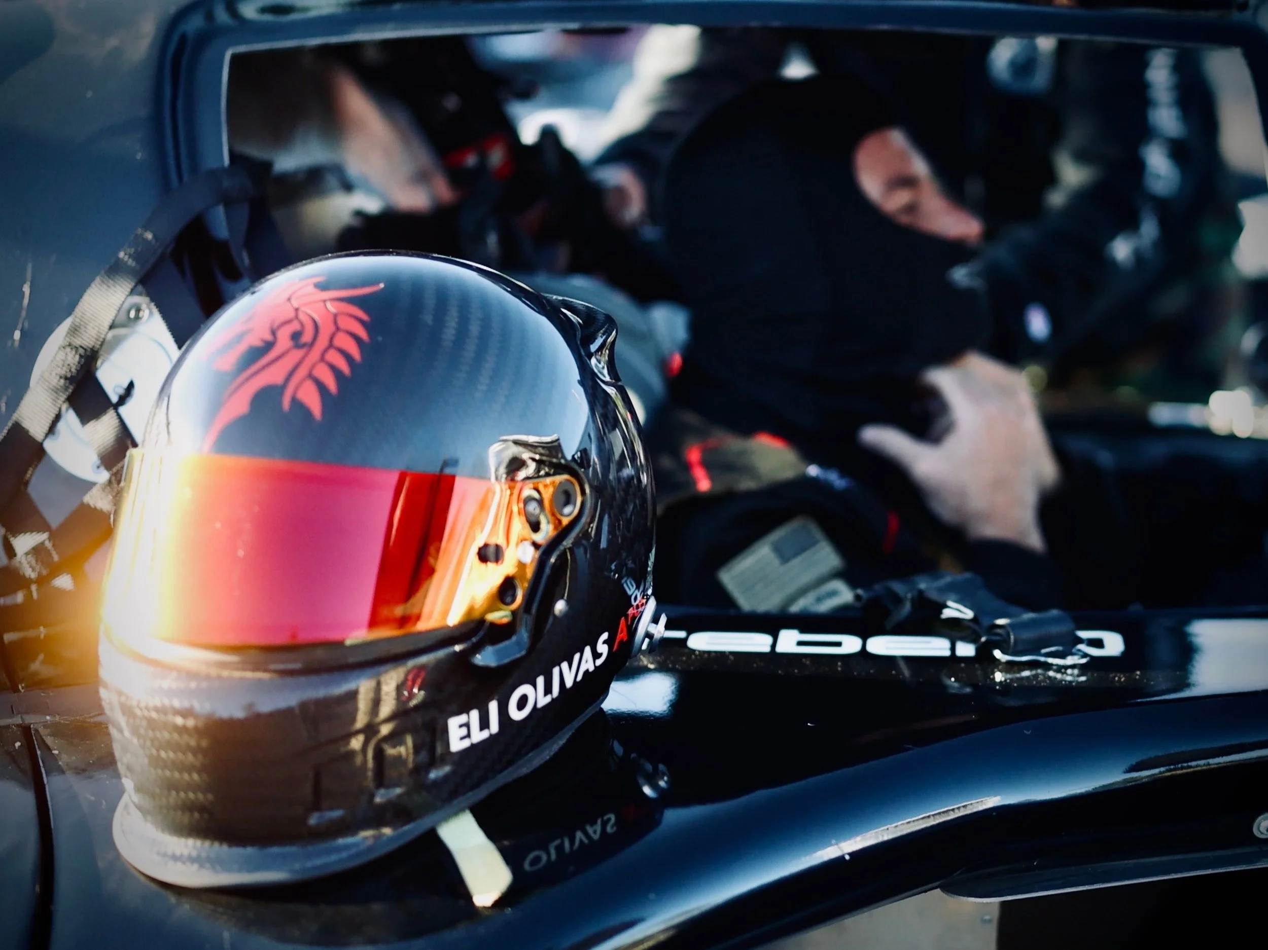 Close-up of a racing helmet with a red dragon logo, reflected on a shiny black surface, with a blurred background showing people in racing gear.