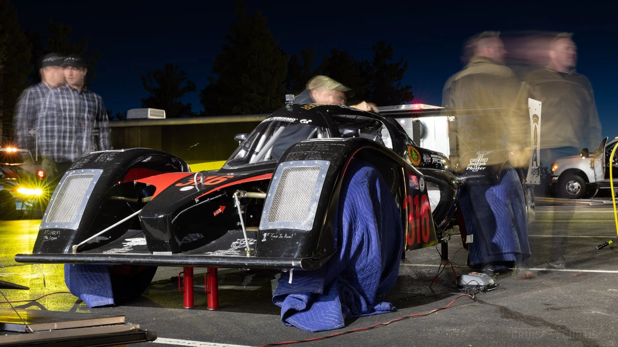 A race car is being worked on at night, with multiple people around it. The car is black with red accents and has protective blue covers on the wheels. People are blurred due to movement, and equipment and a white vehicle are visible in the backgroun