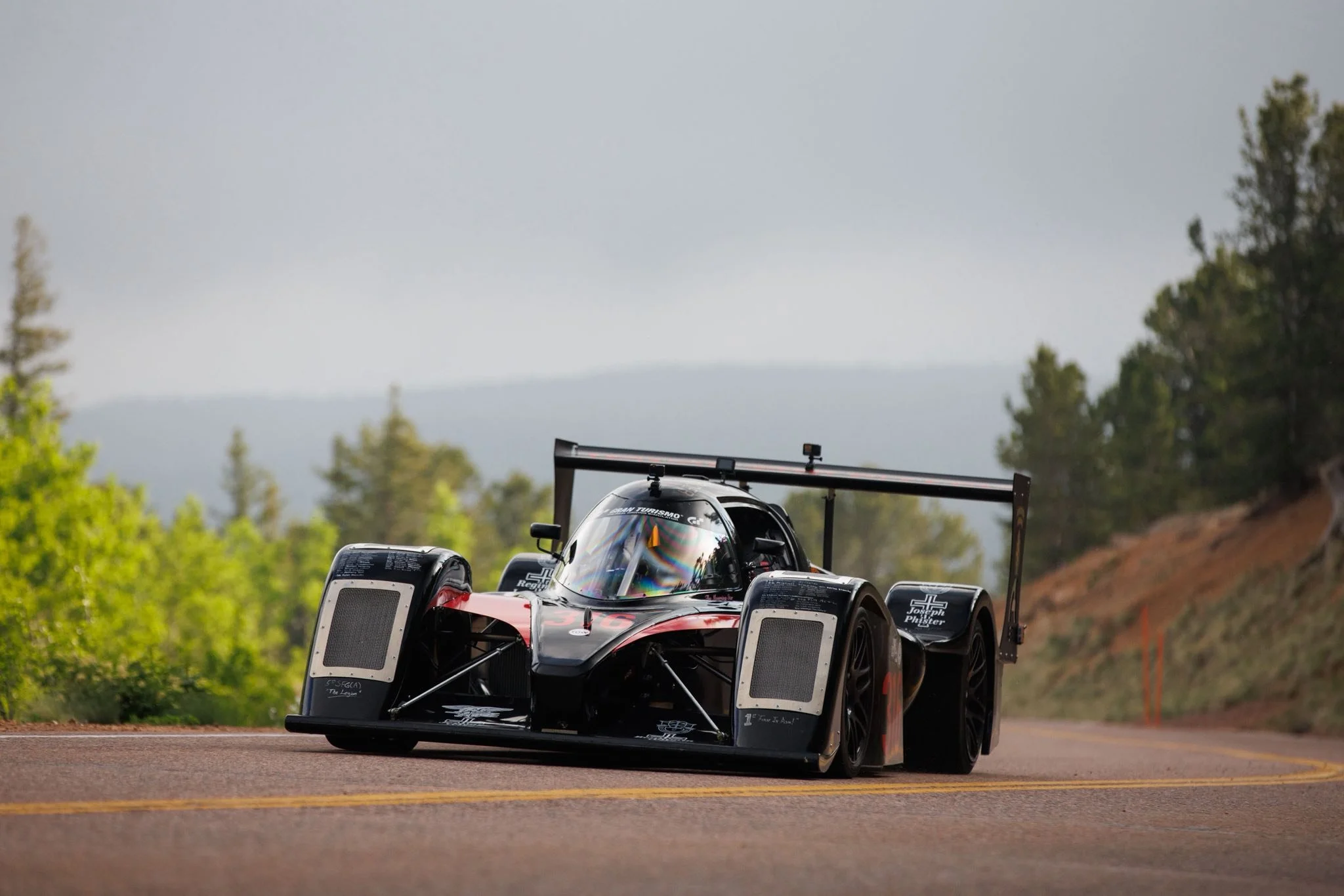 A sleek black race car with aerodynamic design and a rear wing, driving on a paved mountain road surrounded by lush green trees and hillside, with a cloudy sky in the background.