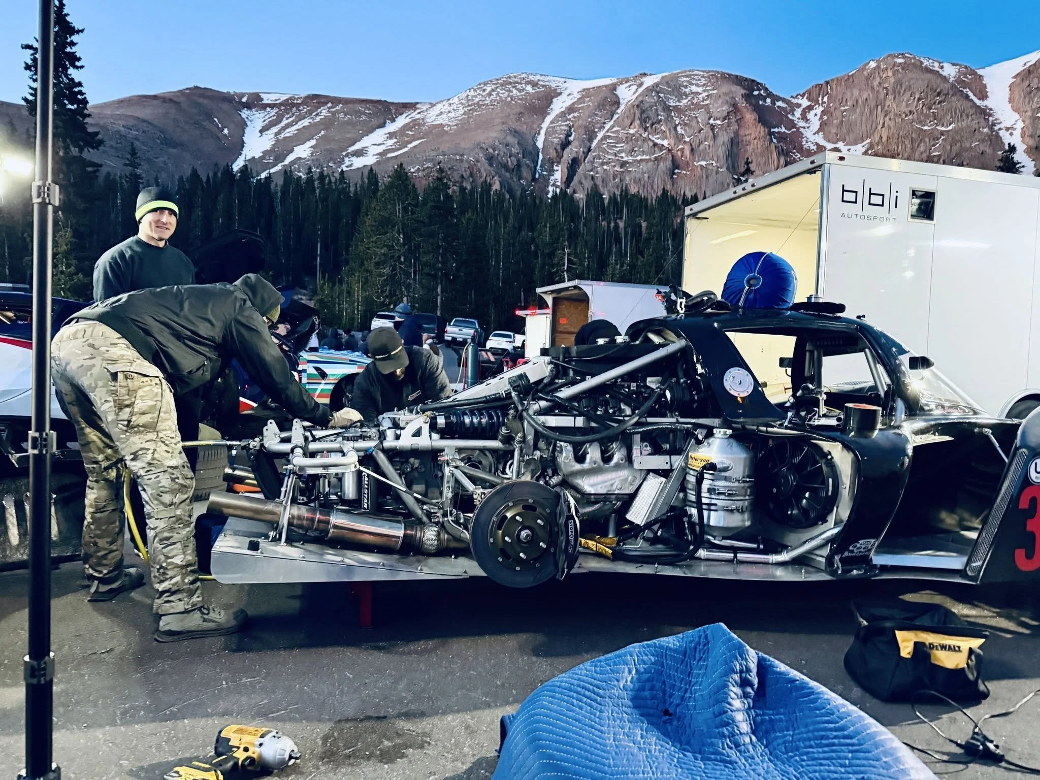 A group of men working on a race car chassis outdoors with mountains in the background. Equipment and tools are scattered around the work area.