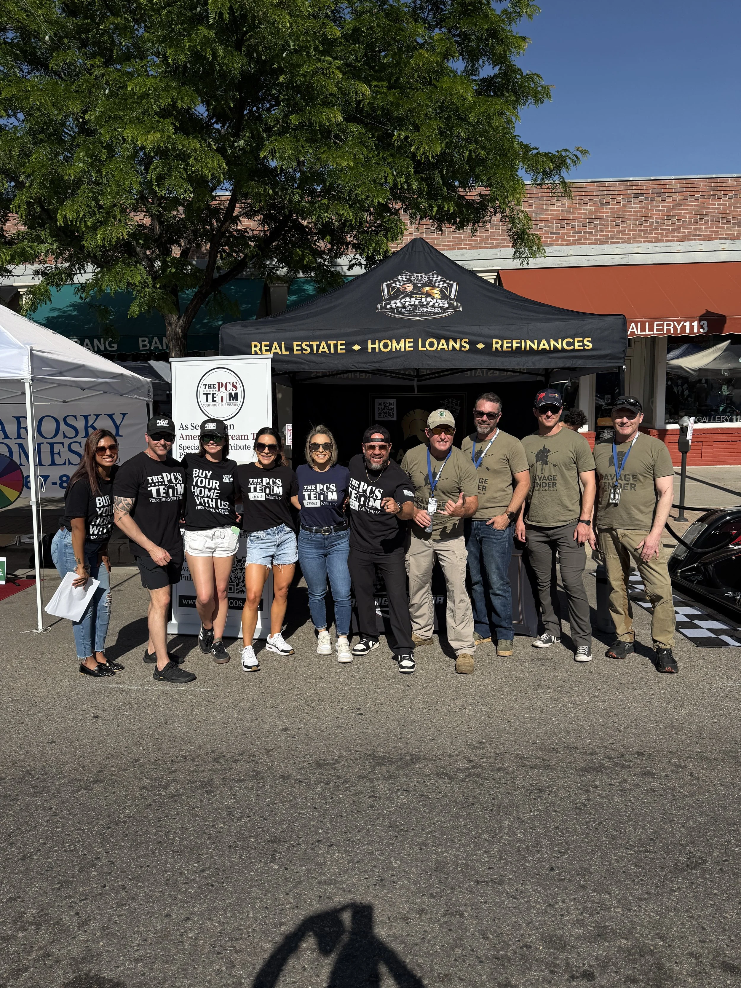 A group of people standing in front of tents at an outdoor event, with a brick building and a tree in the background. The group is smiling and posing for the photo.