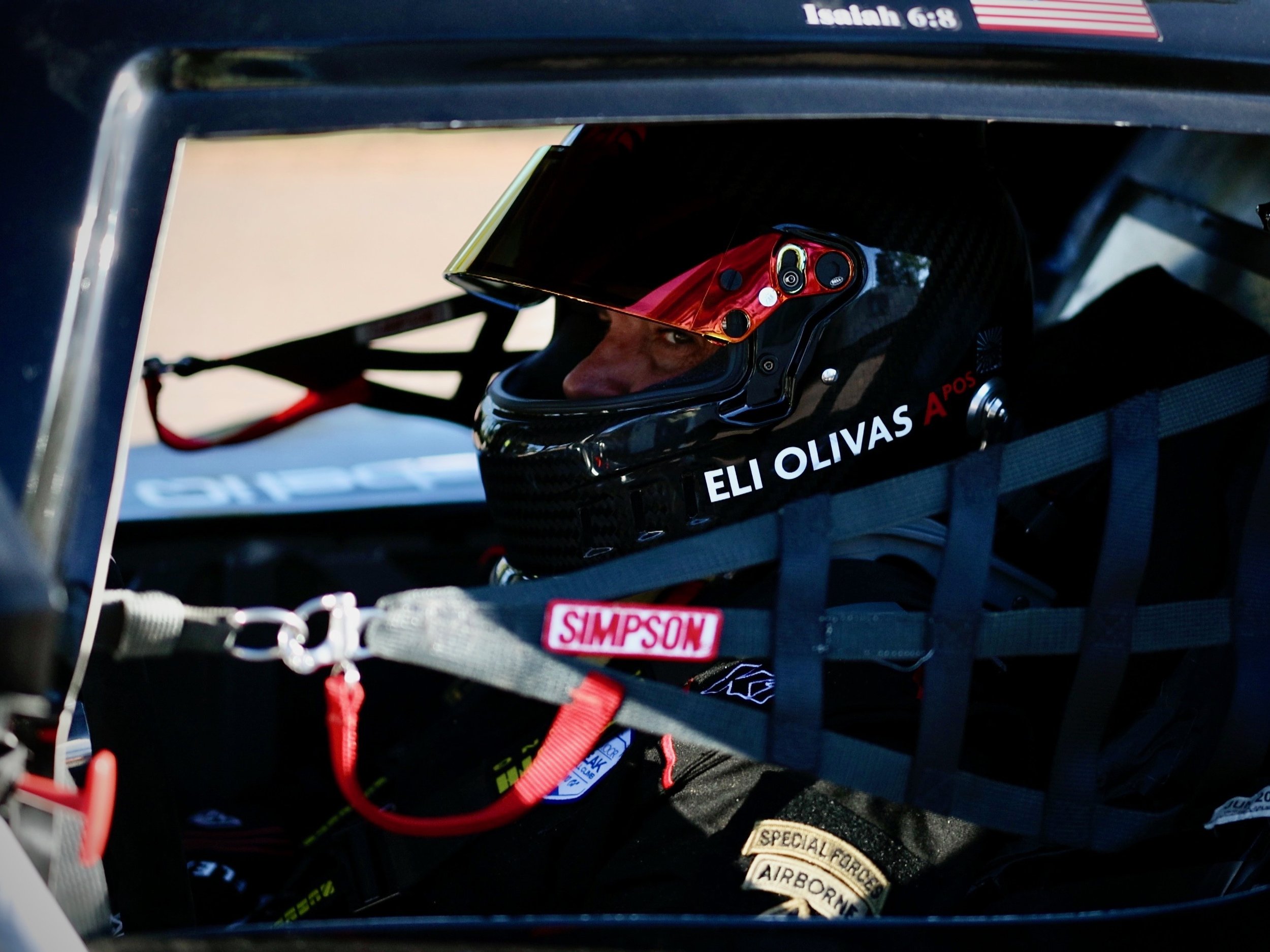 Close-up of a race car driver wearing a helmet with a red visor inside a racing vehicle. The helmet displays the name Eli Olivas. The image reveals parts of the vehicle's interior, including safety harnesses and equipment.