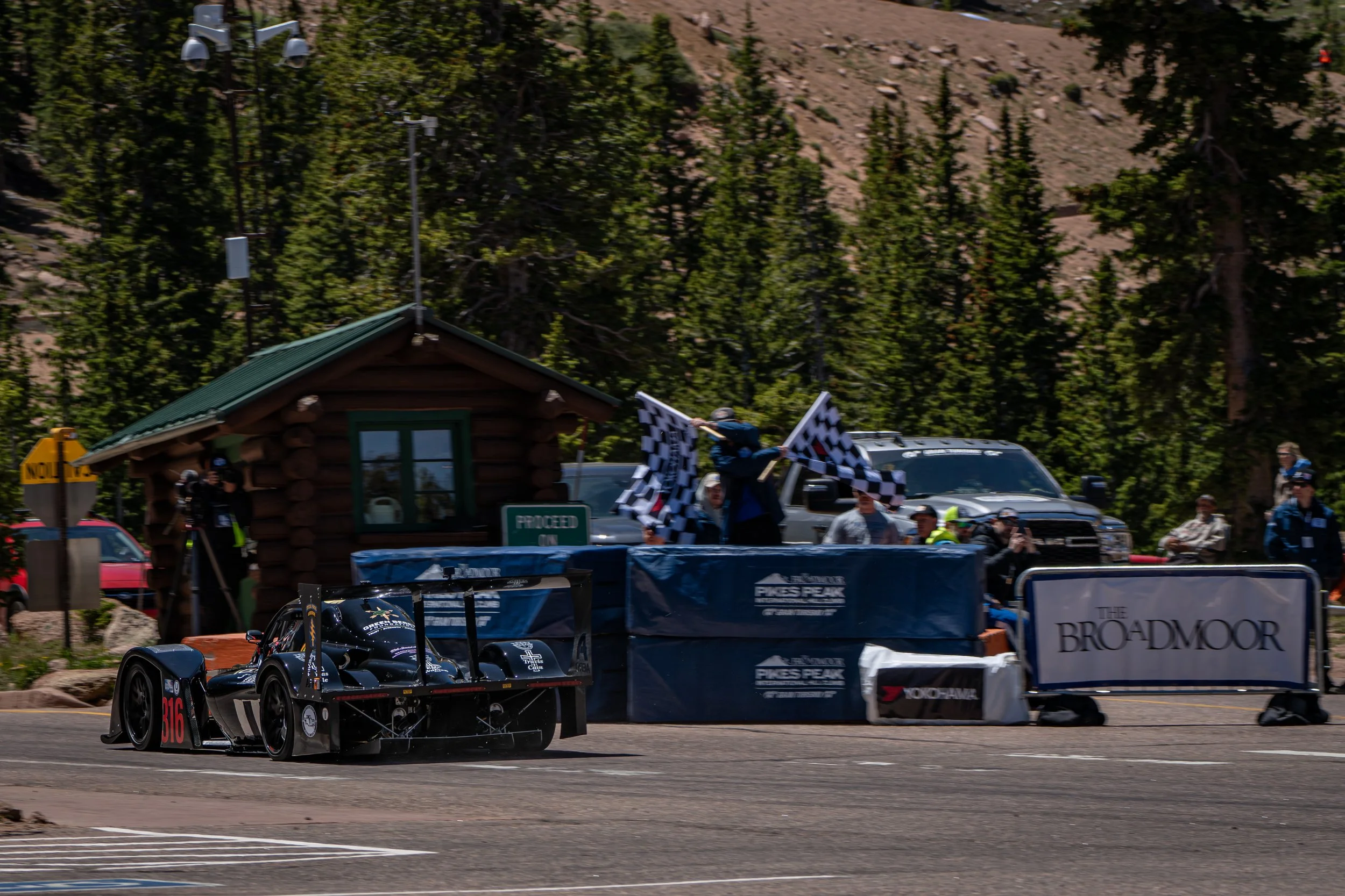 A race car with the number 316 driven by a Formula E driver on a racing track near a wooded area, with a person holding a checkered flag.