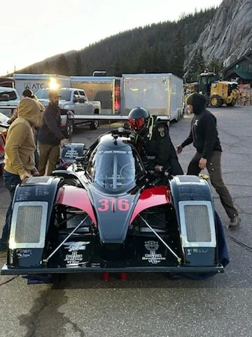 A race car with the number 316 being prepared by a driver in racing gear, surrounded by people and trucks in a mountainous area.