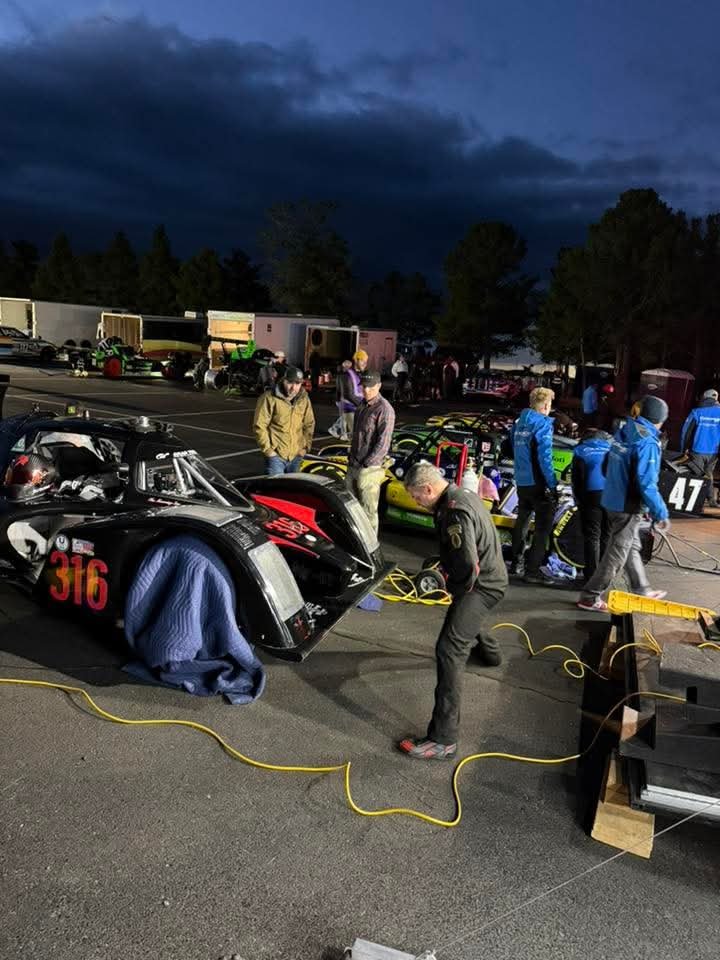 Race cars being prepared and team members working on them in a racing paddock at night under dark clouds.