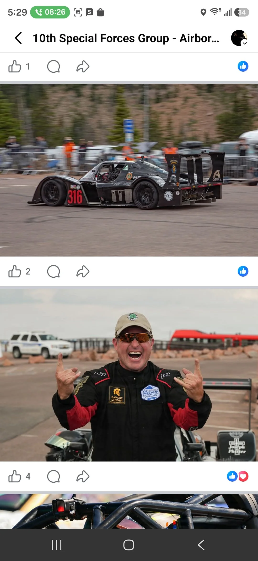 A black race car with number 316 on the front, driving on a race track with people and trees in the background, and a man in racing gear smiling and making the rock on hand gesture.