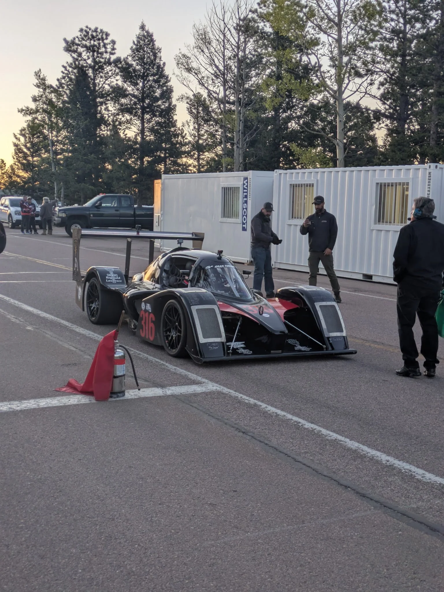 A race car with the number 316 on it parked next to a fire extinguisher and orange caution cone in a parking lot. Several people are standing and talking nearby, and there are large white containers and parked vehicles with trees and a sunset in the 