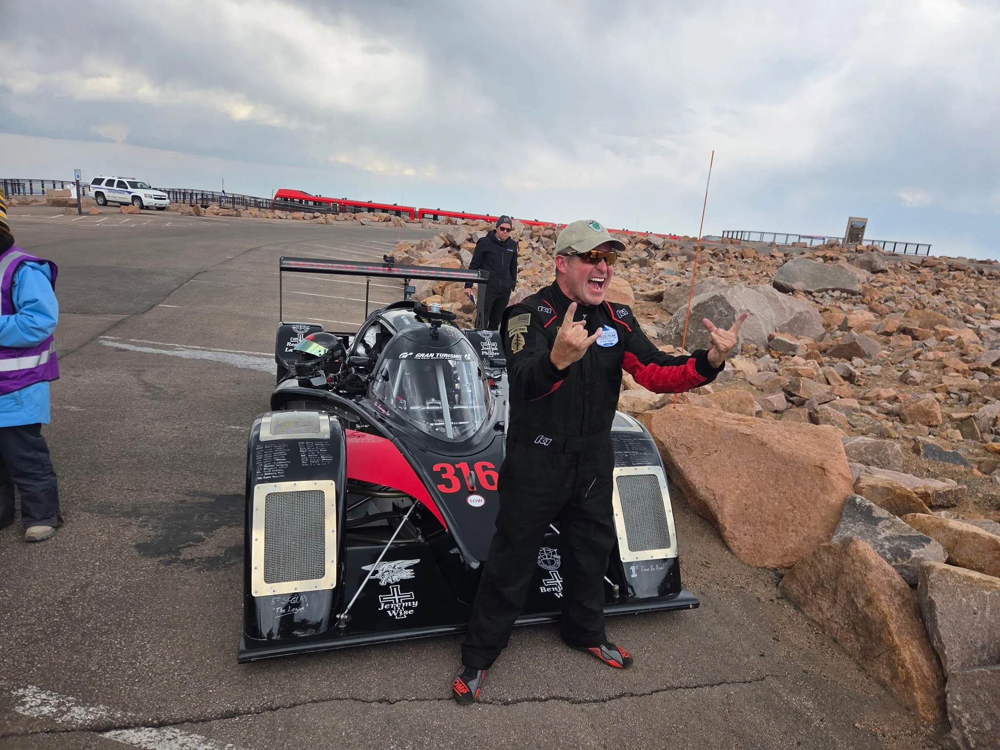 Man in racing suit and sunglasses standing next to a racing car with a group of people and rocky terrain in the background.