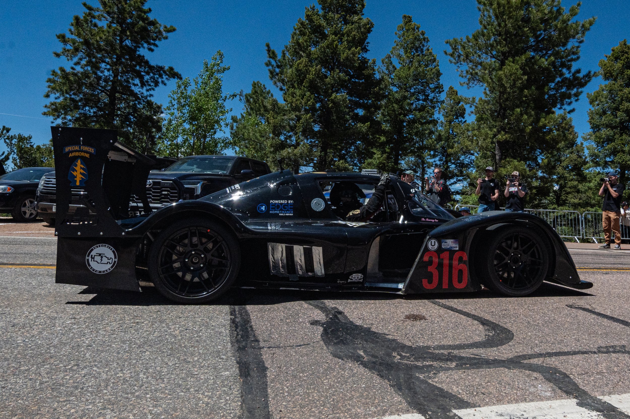 Black race car with racing number 316 parked at an outdoor event, surrounded by people taking photos and videos, with tall green trees and blue sky in the background.