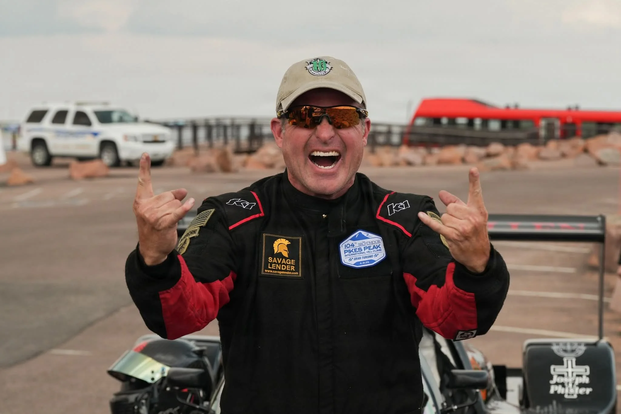A man in racing gear smiling and making a rock and roll hand gesture at a racing event.
