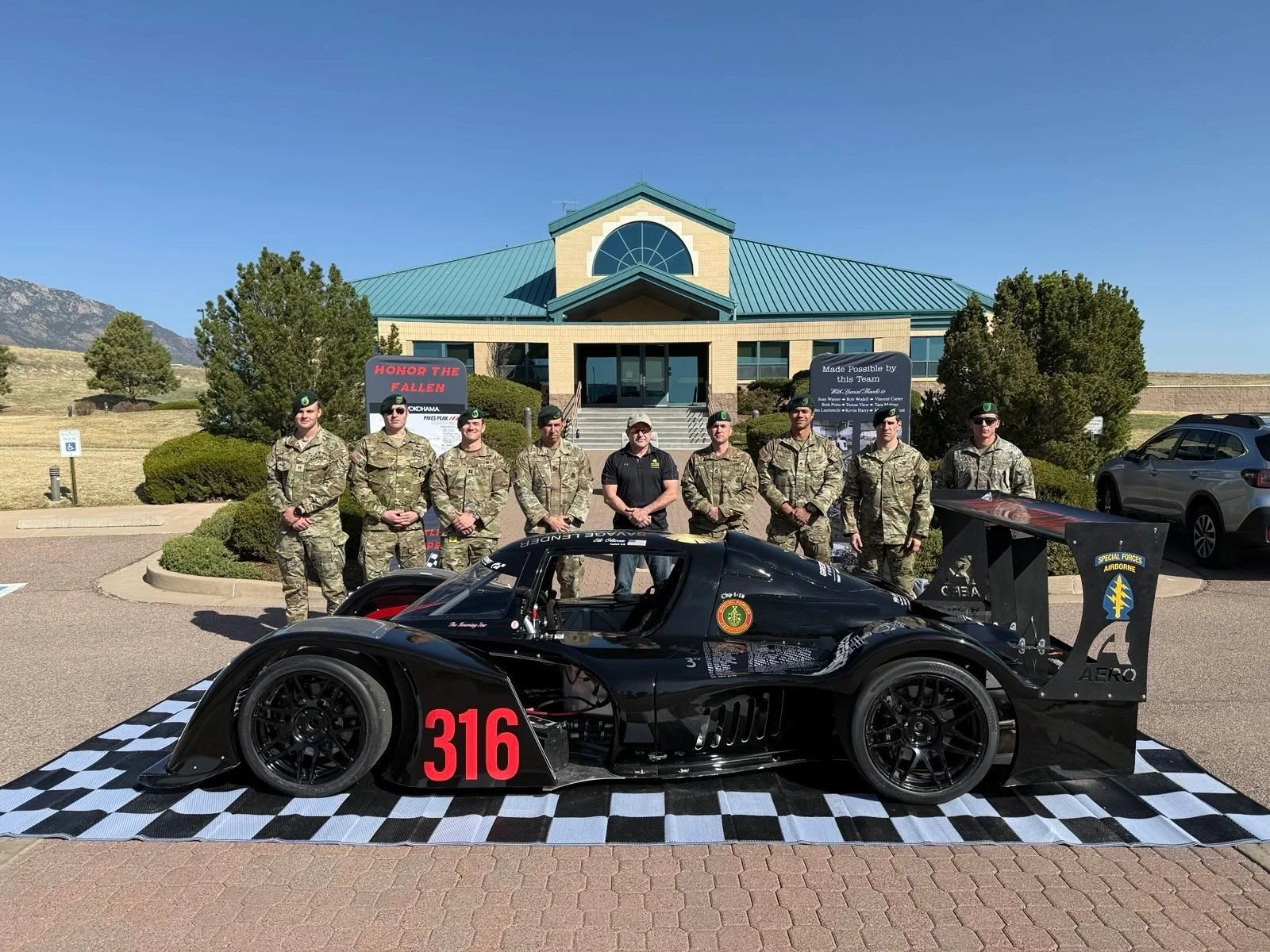 Group of military personnel standing behind a black race car with a checkered flag ground covering, in front of a building with trees and mountains in the background.