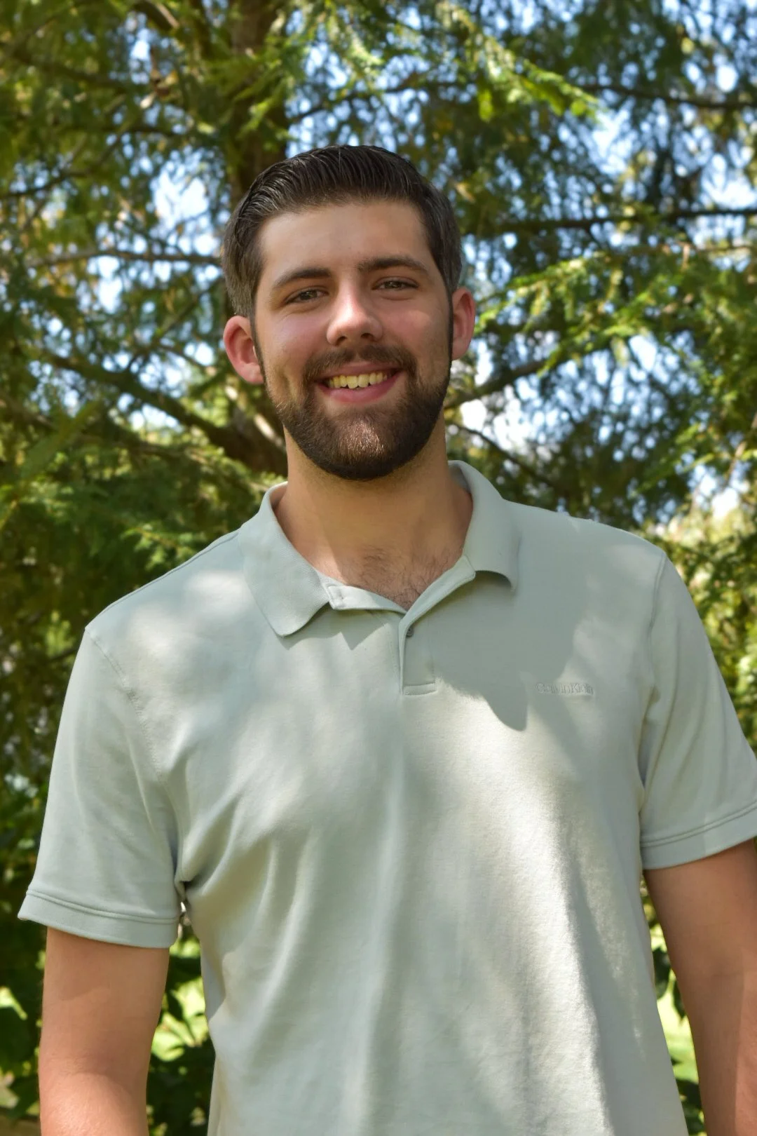 A young man with dark hair and a beard smiling outdoors in front of a leafy tree.