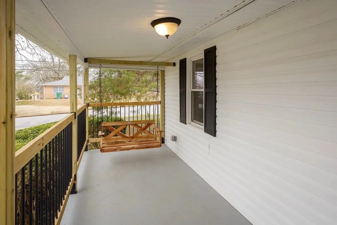 Empty front porch with a wooden swing hanging from the ceiling, black railing, and a house with white siding and black shutters in the background.