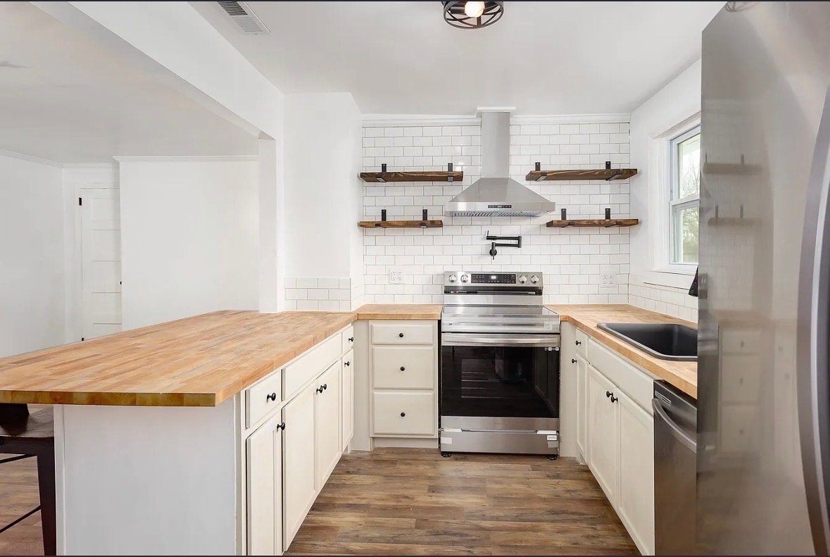 Minimalist kitchen with white cabinets, wooden countertops, stainless steel oven, range hood, window, and open wooden shelves on white tiled wall.