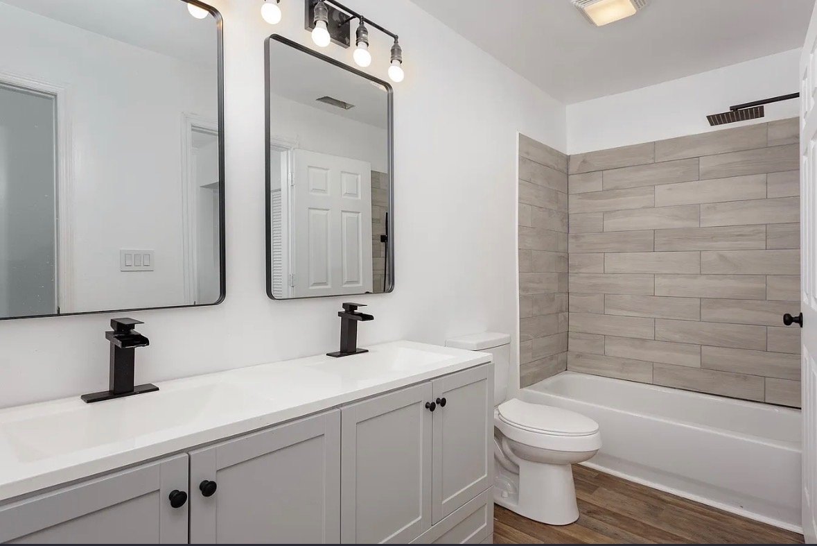 Modern bathroom with double vanity sinks, large mirrors, black fixtures, a toilet, and a bathtub with light gray wood-like tiles on the wall.