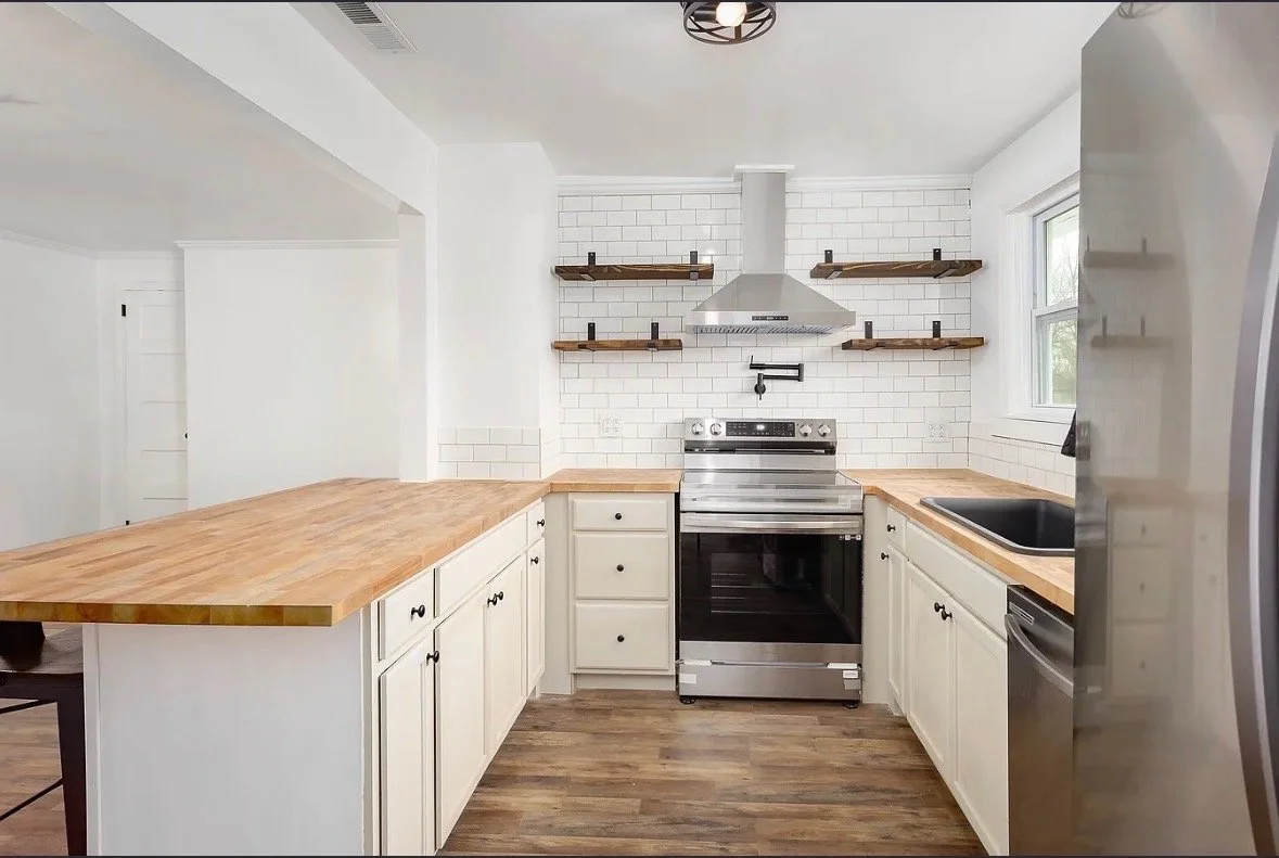 Modern kitchen with white cabinets, stainless steel appliances, wooden countertops, white subway tile backsplash, open floating wooden shelves, and a window.