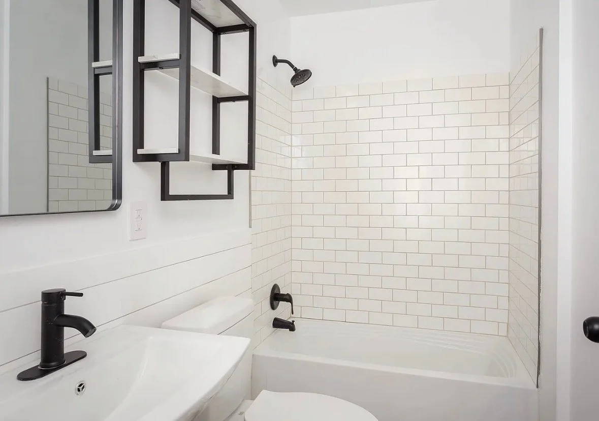Modern bathroom featuring a black showerhead, white subway tile walls in the shower area, a white bathtub, a white vanity with a black faucet, a unique black metal wall shelf, and a mirror reflecting part of the room.