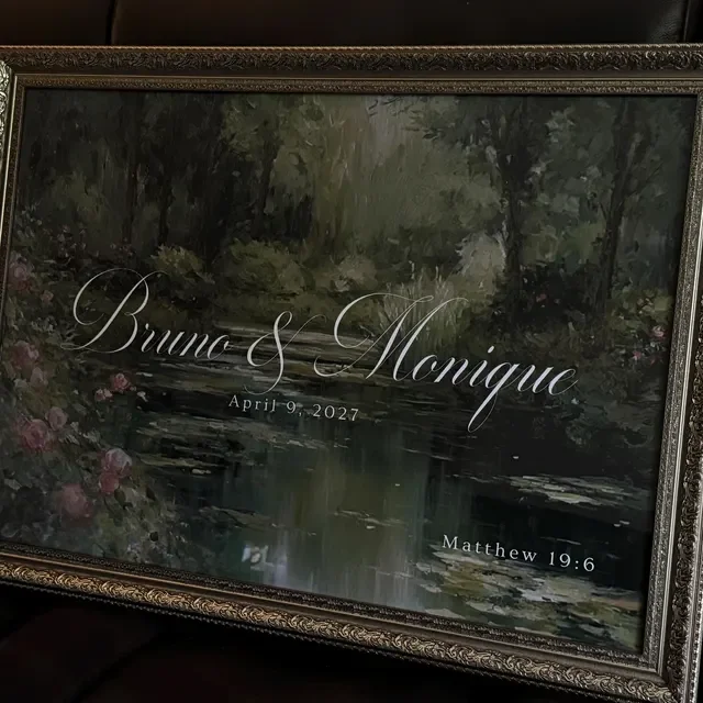 A framed wedding welcome sign featuring a painted peaceful river scene surrounded by greenery, with elegant text overlay reading "Brune & Monique," the date "April 9, 2027," and a Bible verse reference "Matthew 19:6." Handmade by January Swift Studio