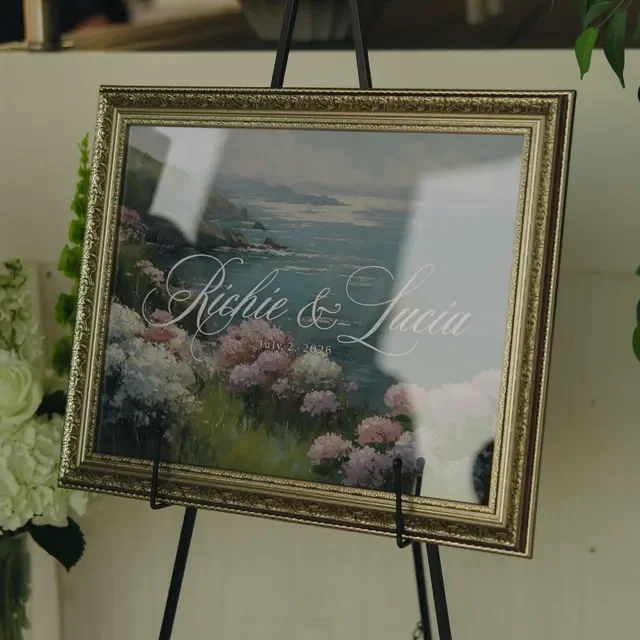 Framed wedding sign on easel with the names Richie and Lucia, and a date July 2, 2026, with a landscape painting featuring flowers and ocean in background.  Handmade by January Swift Studio.