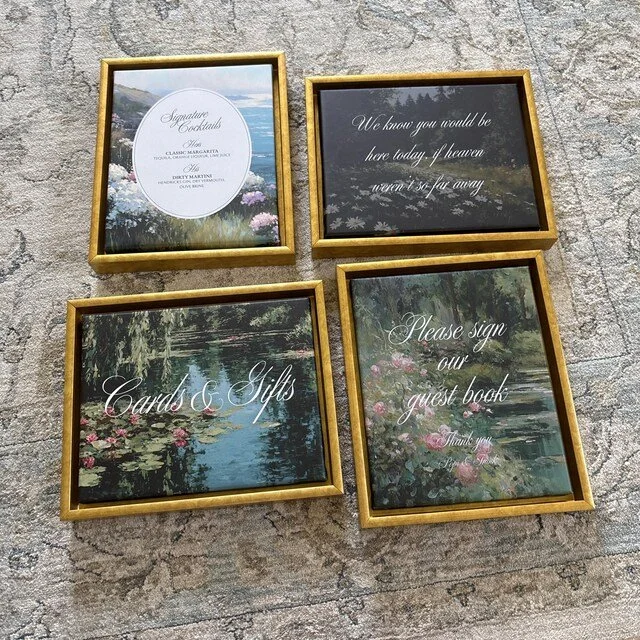 Four decorative wedding signs in gold frames with nature scenes and calligraphy text, including a menu, a quote, and messages for cards, gifts, and a guest book, arranged on a stone surface. Handmade by January Swift Studio.
