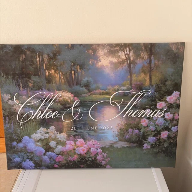 A wedding welcome sign with a painted landscape background of trees, flowers, and a river, displaying the names "Chloe & Thomas" and the date "26th June 2026" in elegant script. Handmade by January Swift Studio.