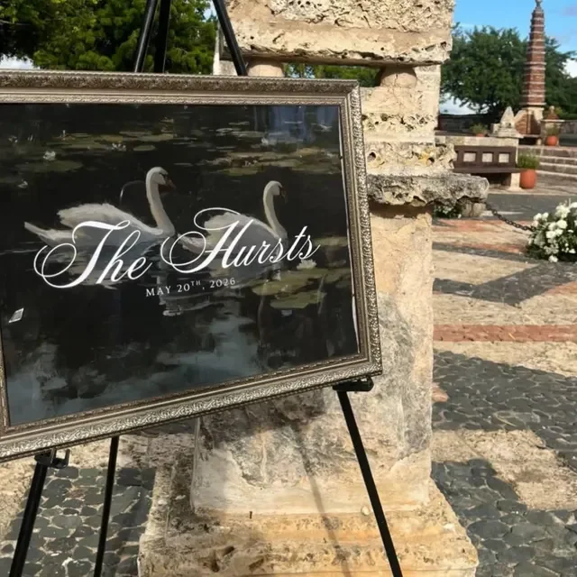 A wedding welcome sign featuring two swans on a pond, with the text 'The Hursts' and a date 'May 20th, 2026', leaning against a stone wall outdoors. Handmade by January Swift Studio.