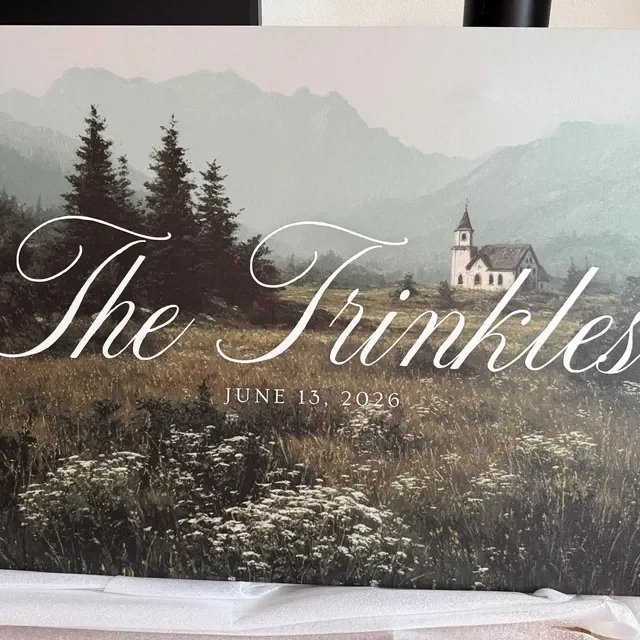 A wedding welcome sign featuring a painted scenic landscape with a grassy field, wildflowers, trees, and mountains in the background. There is a small white church with a steeple in the distance. The overlay text reads "The Trinkles" with the date Ju