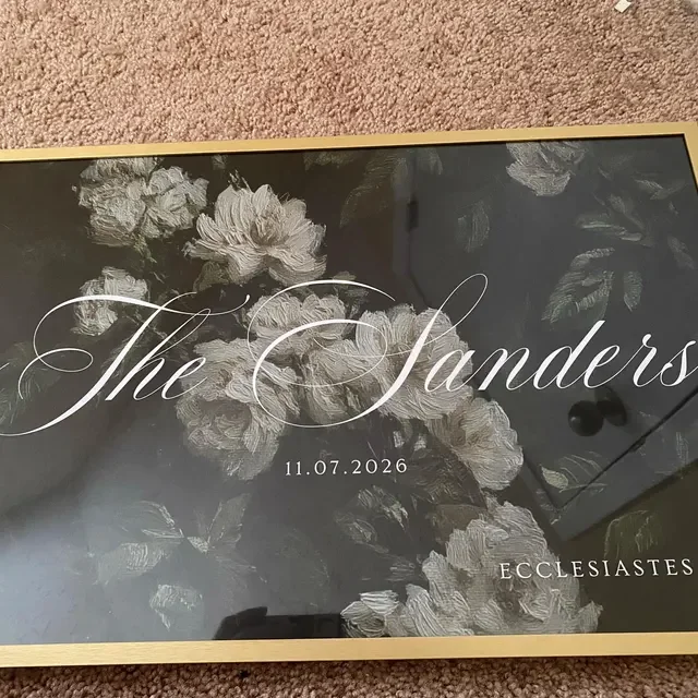A black and gold wedding sign with a painted floral design, featuring the names 'The Sanders', a date '11.07.2026', and the word 'Ecclesiastes'. Handmade by January Swift Studio.