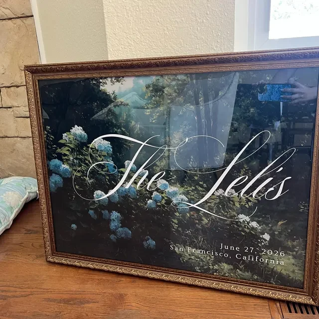 Framed wedding welcome sign with a painted floral background and elegant cursive text reading "The Lex," with additional text indicating a date of June 27, 2026, and location San Francisco, California. Handmade by January Swift Studio.