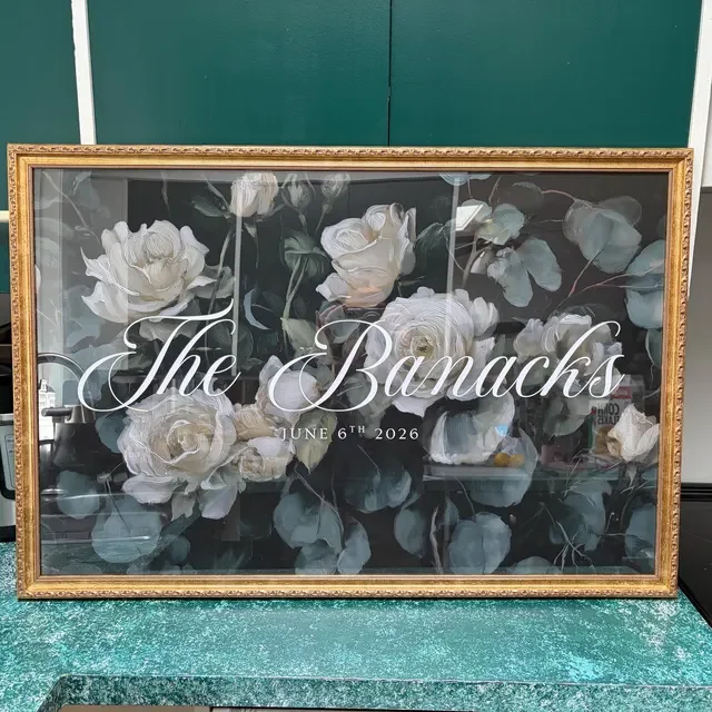 Framed wedding welcome sign featuring white roses and green foliage background. Elegant script text reads "The Banachs June 6th 2026." Handmade by January Swift Studio.
