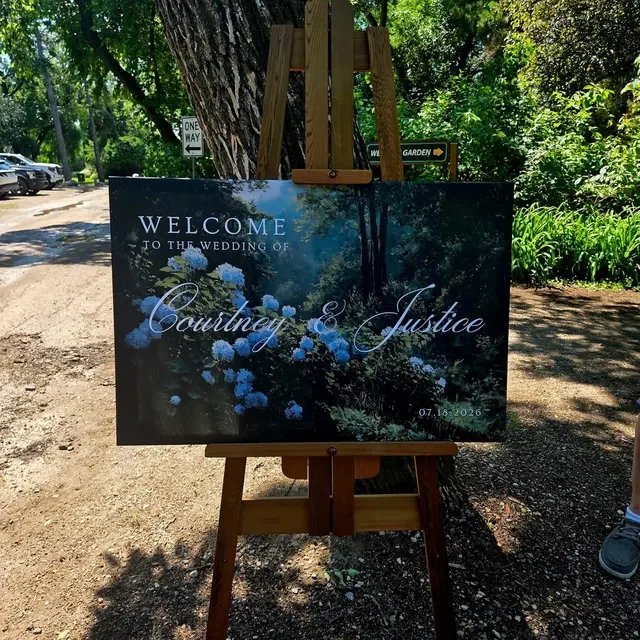 Welcome sign for the wedding of Courtney and Justice, dated July 18, 2026, with a background of white flowers and greenery. Handmade by January Swift Studio.