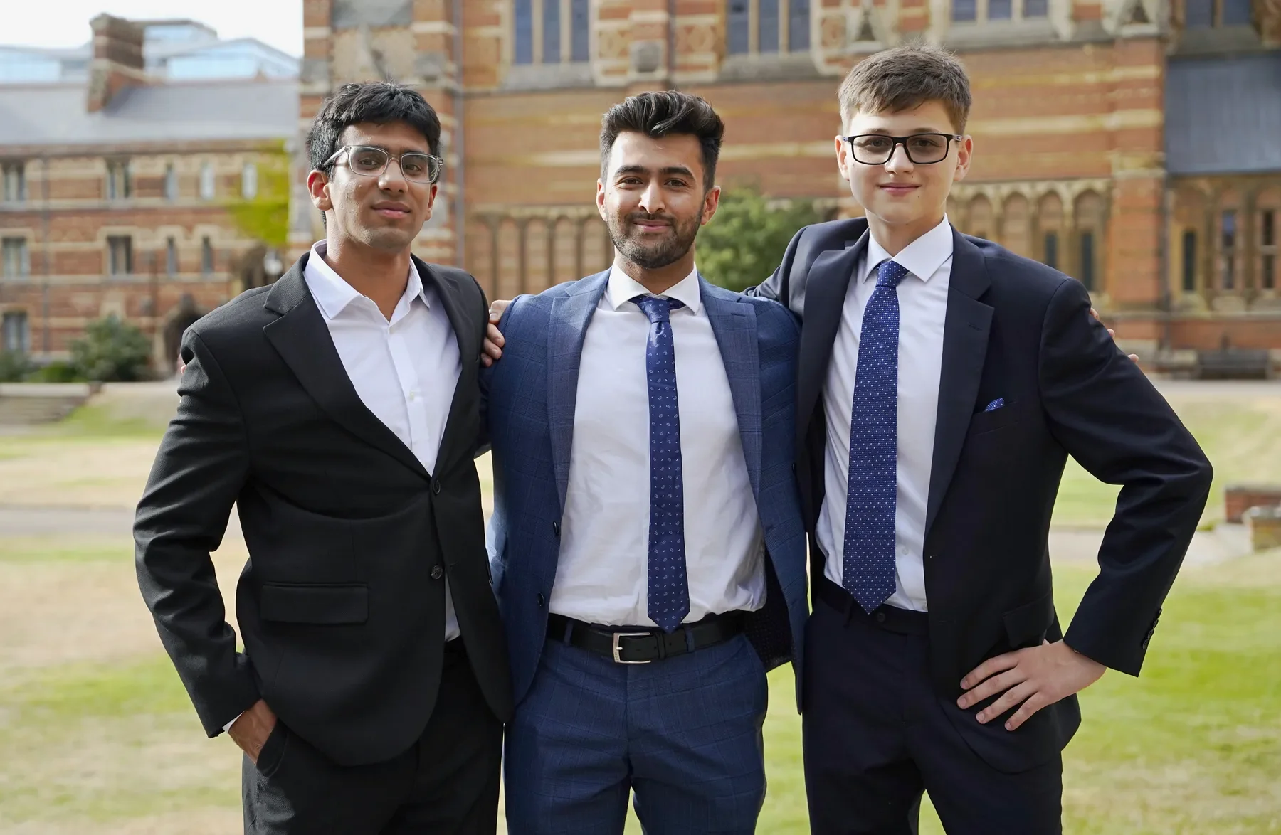 Three young men in suits standing outdoors in front of a historic brick building, smiling and posing with their arms around each other.