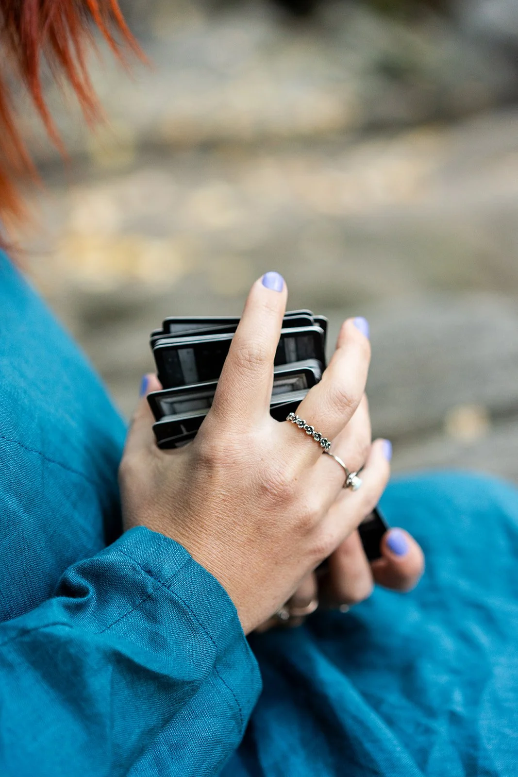 A person with pink hair, wearing a blue jacket, sitting outdoors, holding a handful of small black and white cameras or devices.