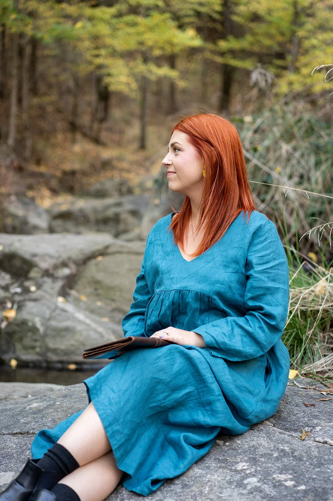 A woman with red hair wearing a blue dress sits on a rock near a creek in a wooded area with autumn foliage.