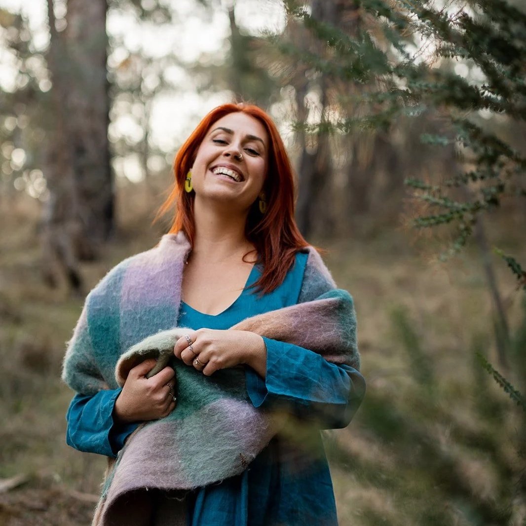 A woman with red hair smiling outdoors in a forest, wrapped in a multicolored shawl over a blue dress.