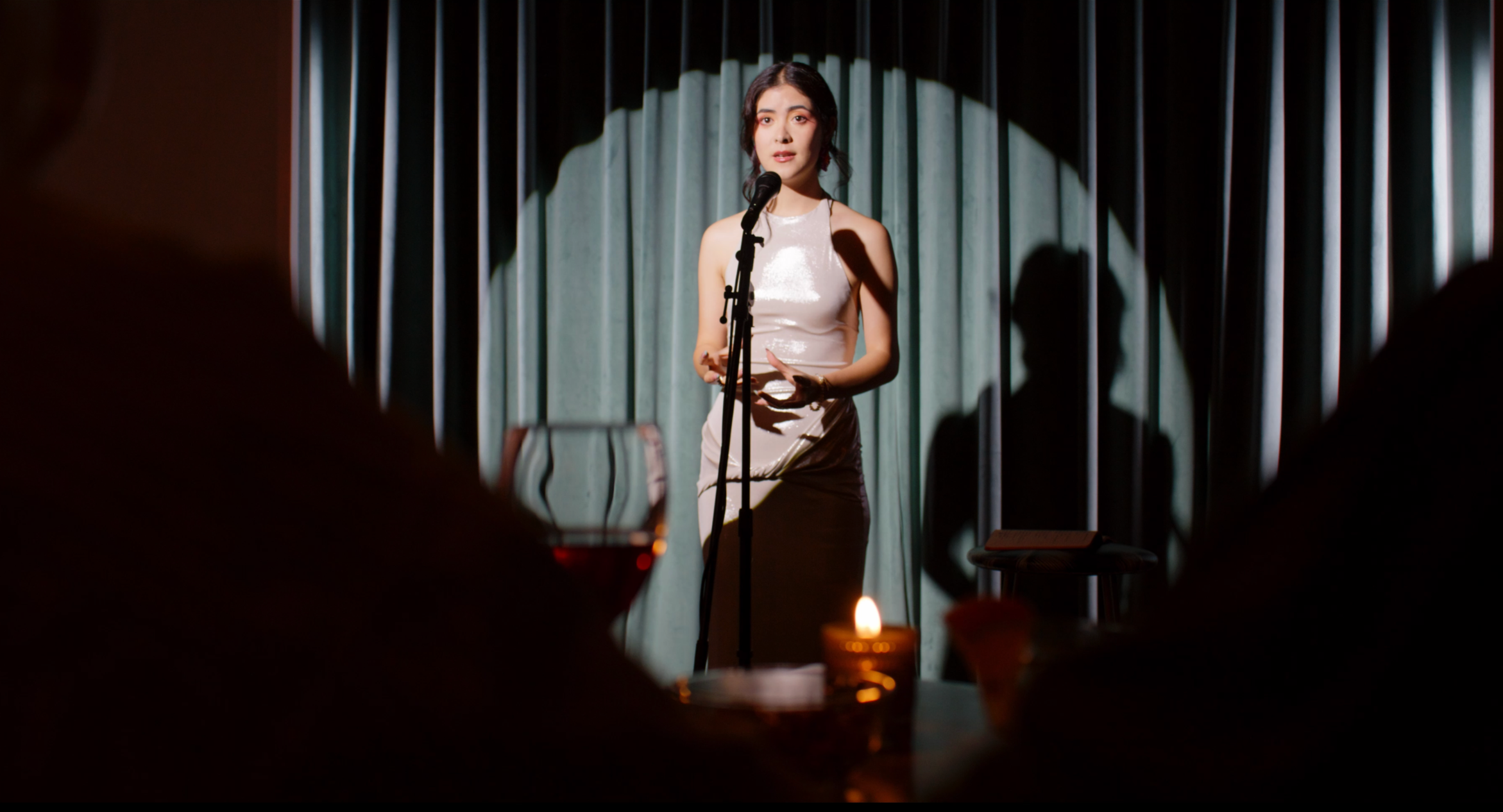 A woman in a shiny sleeveless dress is speaking into a microphone on a stage with teal curtains. The scene is viewed through an audience, with some silhouette shadows and a lit candle in the foreground.